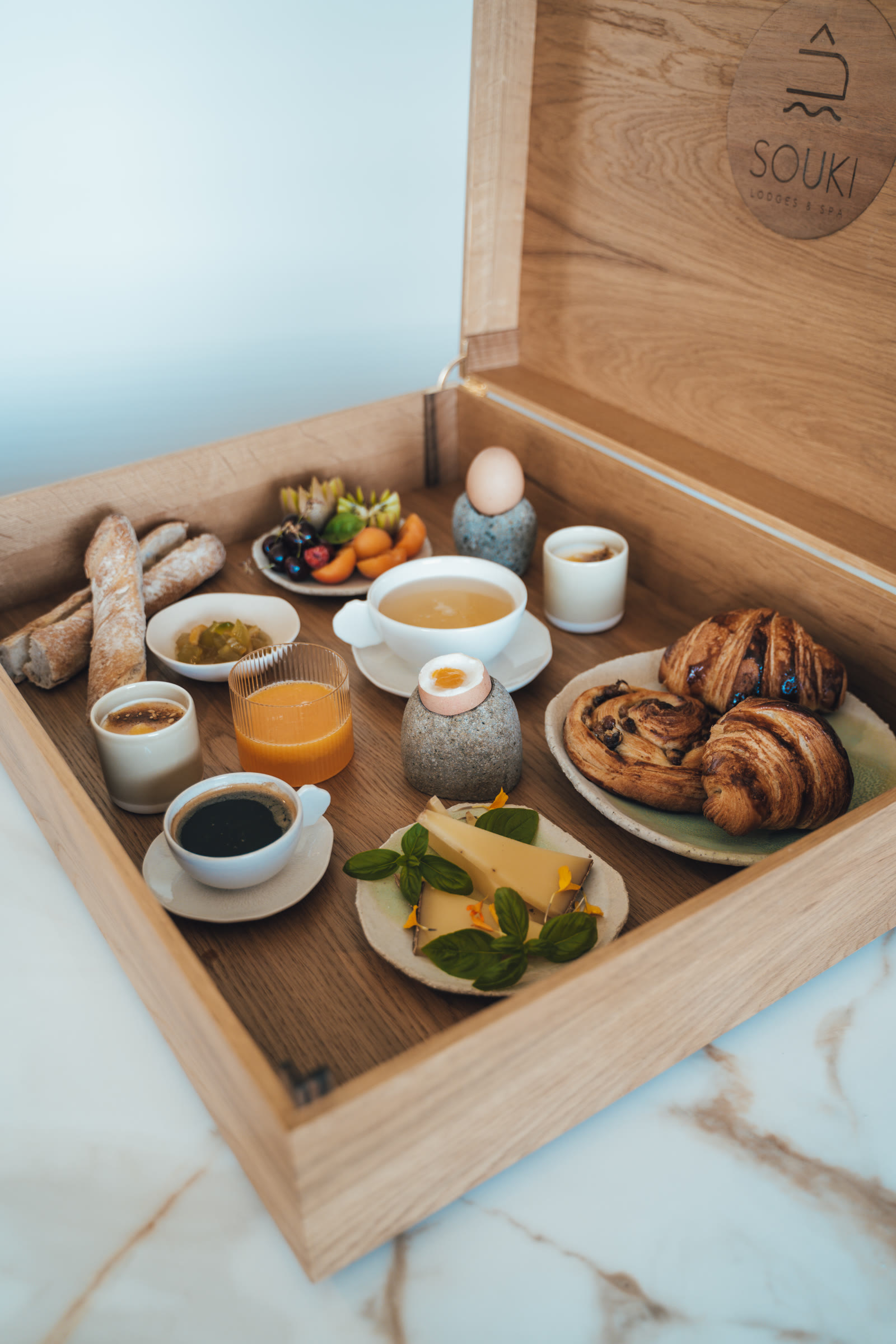 La malle du petit-déjeuner de Souki ouverte, emblème gravé sur le couvercle, viennoiseries, œuf, fruits et jus