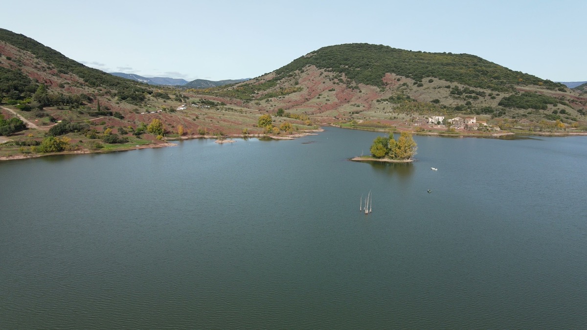 Le lac du Salagou vu du ciel, son îlot boisé et le hameau des Vailhés au pied des pentes rouges