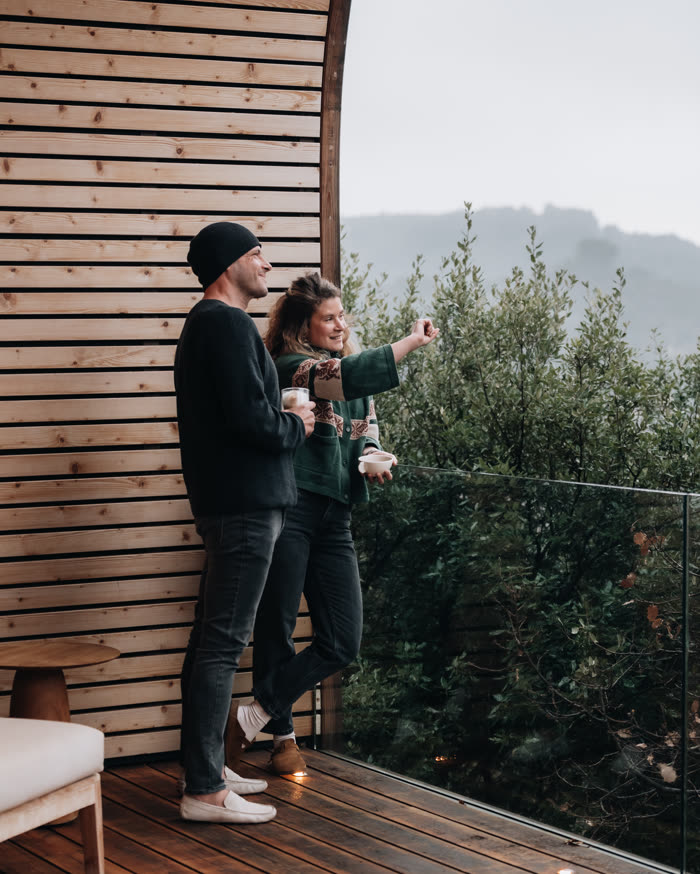 Deux voyageurs debout sur la terrasse d’un lodge, un verre à la main, devant la vallée dans la brume