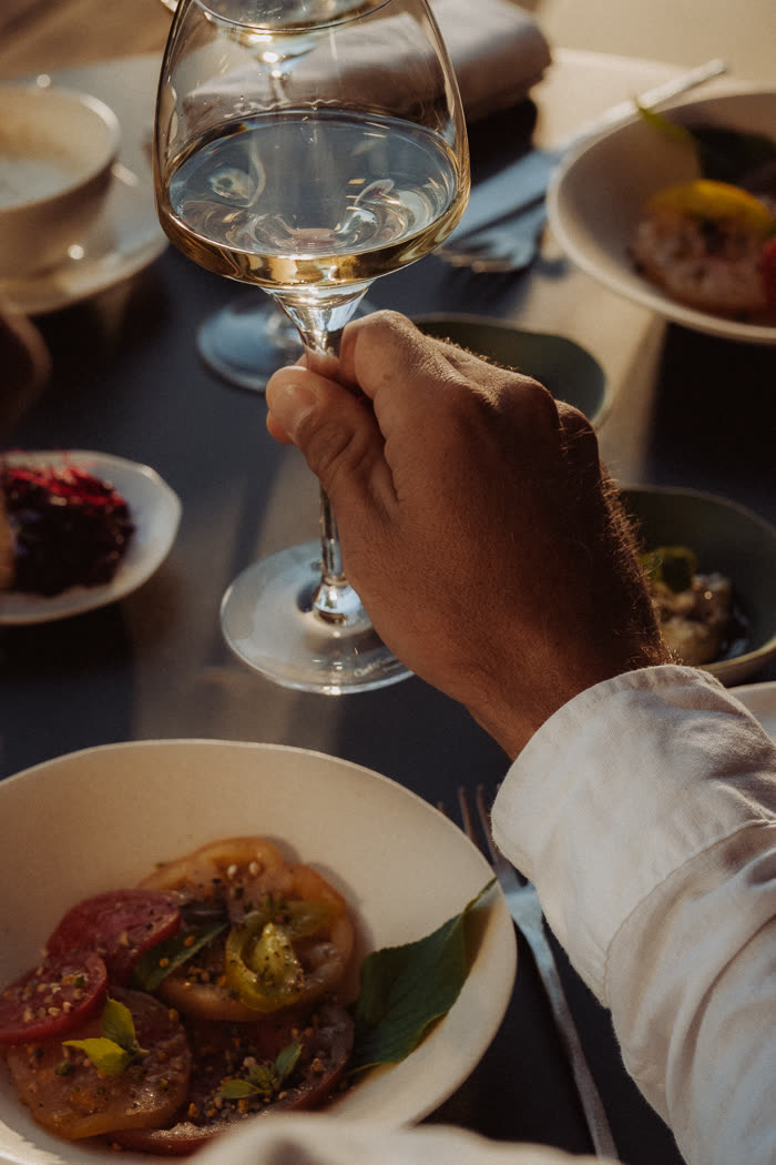 Un verre de vin blanc levé au-dessus d’une assiette de tomates du potager, parmi les entrées du Menu Balade