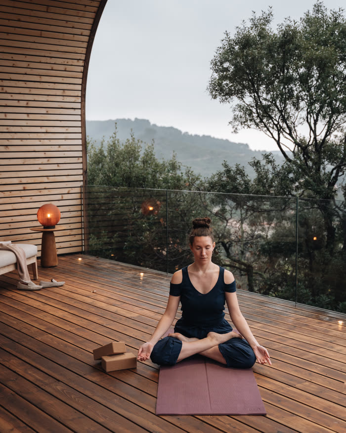 Une voyageuse assise en tailleur sur son tapis, sur la terrasse du lodge, face aux collines