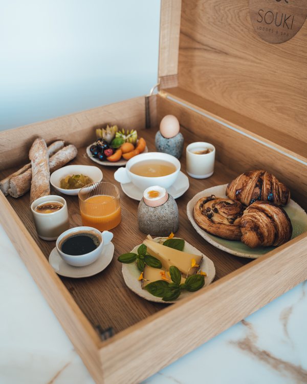 La malle en bois du petit-déjeuner ouverte : baguette, viennoiseries, œuf, jus d’orange et café