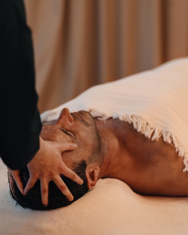 Les mains de la praticienne sur le visage et le crâne d’un voyageur allongé, dans le lodge