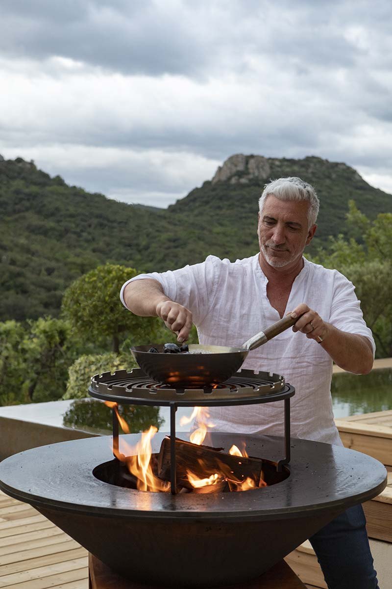 Gilles cooks in the brazier on the terrace, live flames, Cabrières hills in the background