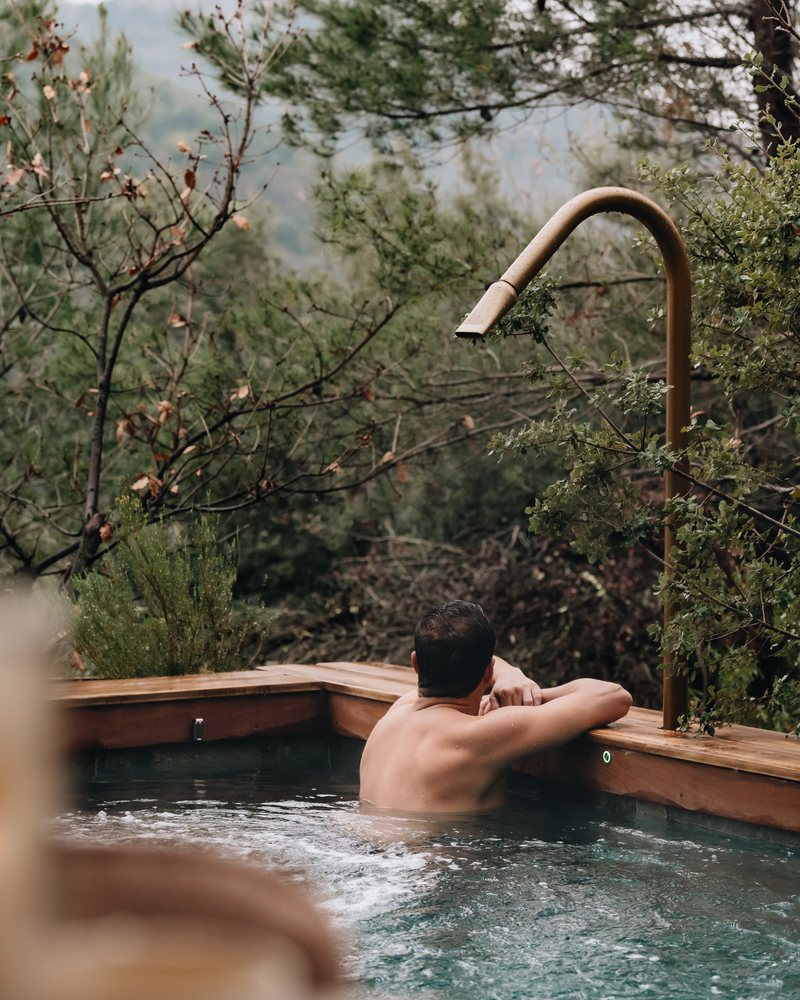Un baigneur accoudé à la margelle de bois de sa piscine privée chauffée, plage à bulles en marche, arbres d’hiver