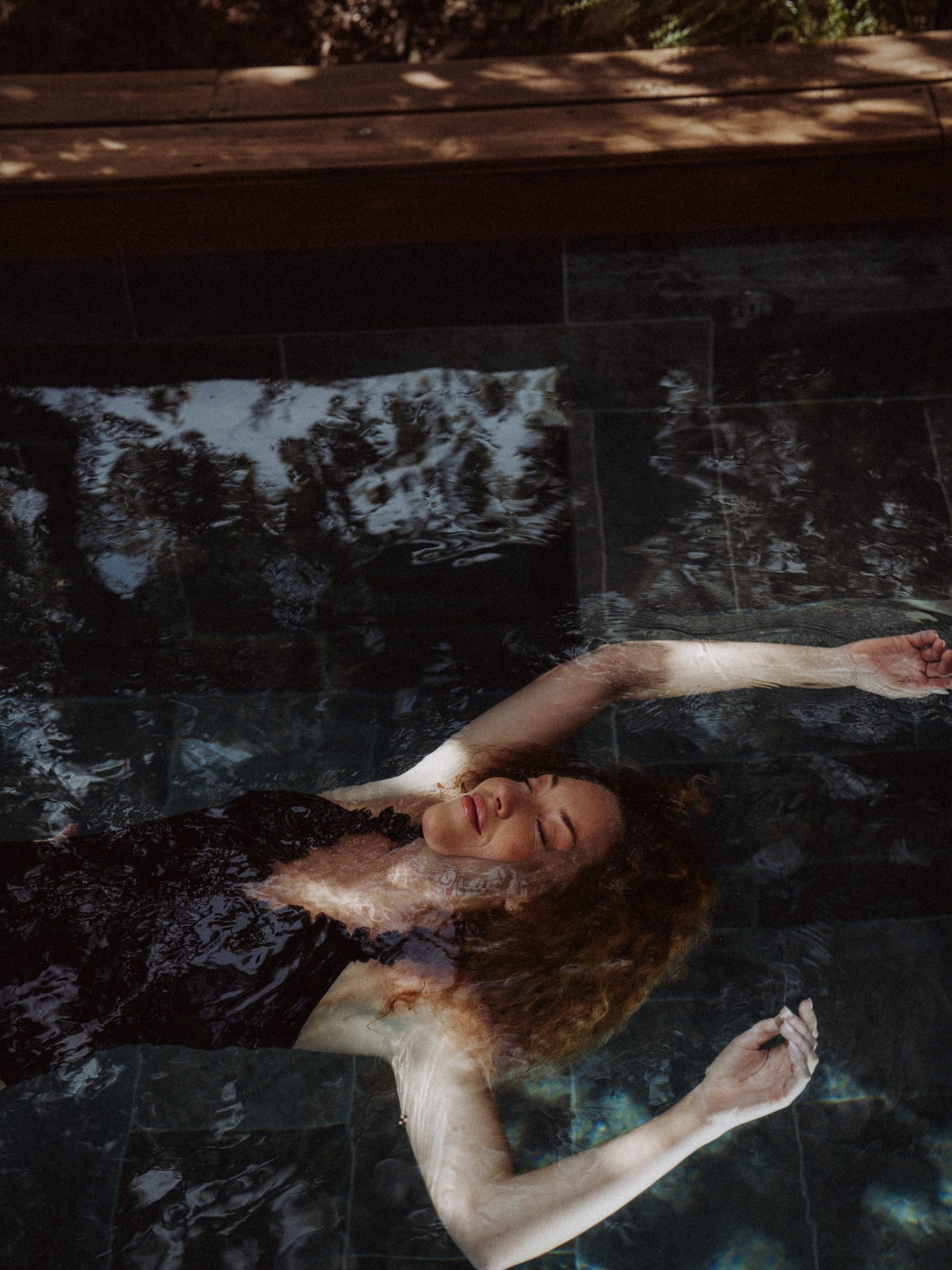 The body weightless at the surface of the pool, arms outstretched, under the reflections of the trees