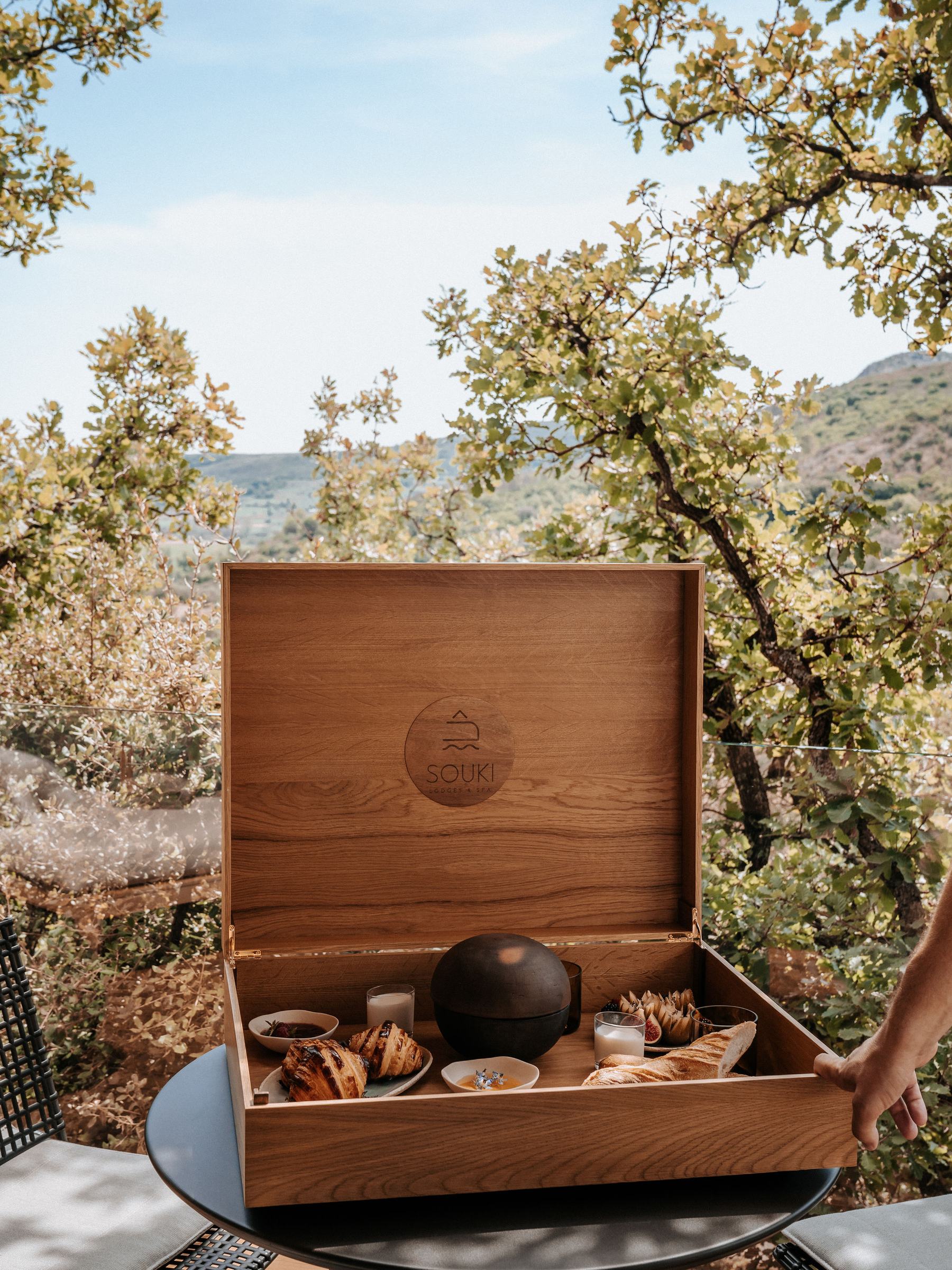 The large wooden chest open, its lid engraved Souki: pastries, bread, jams, figs and yoghurts, outdoors under the oaks